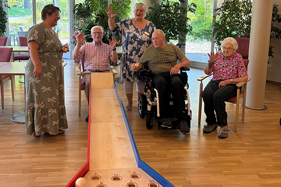 Fünfköpfige Gruppe aus Senior:innen und Betreuung mit der neuen Sitz-Kegelbahn, die auf dem Schoß eines Seniors aufliegt