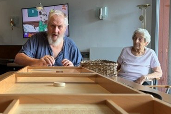 Senior sitzt an Tisch und spielt mit Holz-Scheiben an Holz-Klappspiel. Daneben sitzt eine Seniorin und schaut zu.